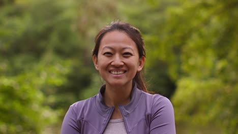 Portrait-Of-Smiling-Mid-Adult-Woman-Exercising-Doing-Work-Out-Outdoors-In-Forest-Wearing-Sports-Clothing